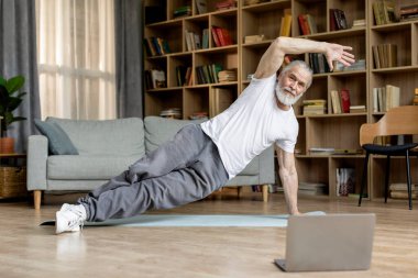 Healthy handsome bearded senior man exercising on yoga mat at home, wearing sportswear, watching fitness video, attending online workout, cozy living room interior, copy space, full length