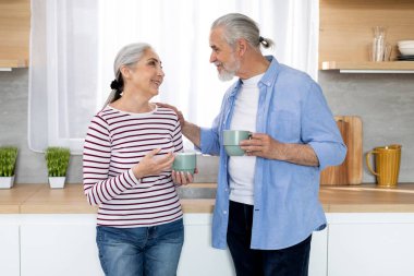 Happy Senior Couple Drinking Morning Coffee And Chatting Together In Kitchen At Home, Loving Elderly Spouses Enjoying Retirement Time, Old Man And Woman Looking At Each Other And Smiling, Copy Space
