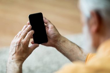 Smartphone with black empty screen in mature man hand, mockup for advertisement. Unrecognizable elderly man using mobile phone, showing nice offer, over shoulder shot, closeup