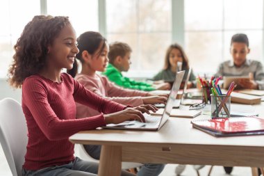Online Learning. Black Schoolgirl Using Laptop Sitting At Desk With Multiethnic Classmates In Classroom Indoor. Modern School Education And Technology Concept. Selective Focus