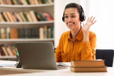 Online tutoring concept. Happy mixed race female tutor having video call with students, waving and talking at laptop webcamera, sitting in library interior
