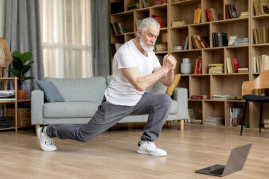 Grey-haired mature man exercising at home, wearing sportswear and sneakers, watching fitness video, attending online workout, cozy living room interior, copy space, full length photo