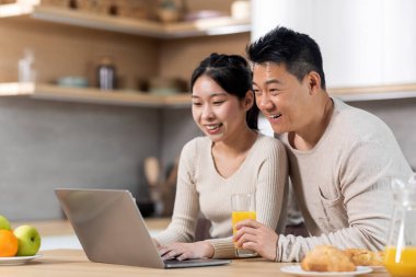 Happy chinese lovers middle aged man and young woman websurfing together while having lunch at home, sitting at kitchen table, drinking fresh orange juice, looking at laptop screen and smiling