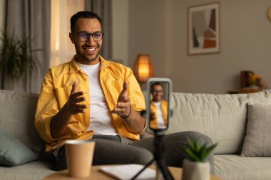 Joyful young African American teacher giving online lesson or lecture, using smartphone at home, copy space. Positive black blogger filming video for social media, live streaming on mobile device