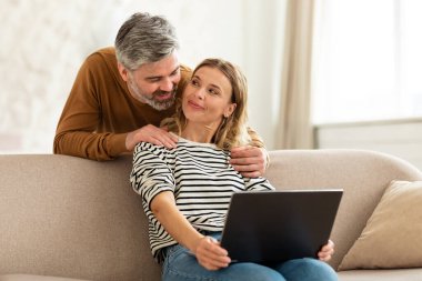 Happy Middle Aged Couple Using Laptop Computer At Home. Husband And Wife Browsing Internet On Computer, Watching Film Online On Weekend. Gadgets And Technology Concept