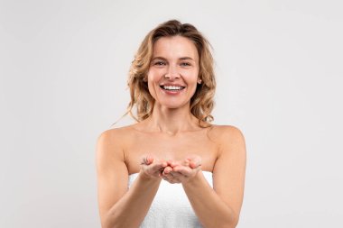 Attractive Middle Aged Female Wrapped In Bath Towel Demonstrating Something On Empty Palms, Beautiful Mature Lady Showing Invisible Object And Smiling At Camera, Posing On Grey Background, Copy Space