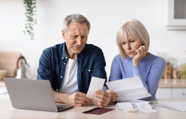 Confused elderly husband and wife checking their monthly expenses, senior man showing his sad wife bill, sitting together in front of laptop at kitchen. Financial hangover for retirees concept