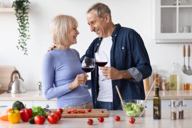 Romantic senior spouses drinking red wine while cooking together at home, loving elderly husband and wife preparing dinner, having conversation, kitchen interior, copy space. Marriage, relationships