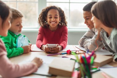 Happy African American Schoolgirl Using Phone Sitting With Classmates At Desk In Modern Classroom Indoors. School Education, Technology And Gadgets. Selective Focus