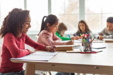 African American School Girl Writing Learning Sitting At Desk In Classroom Indoors. Diverse Children Having Class Taking Notes Indoor. Education And Knowledge. Selective Focus