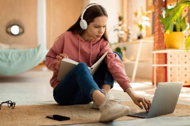 Concentrated teenage girl reading book in front of laptop, getting ready for exam, sitting on floor at home. Focused female student having remote lesson on pc