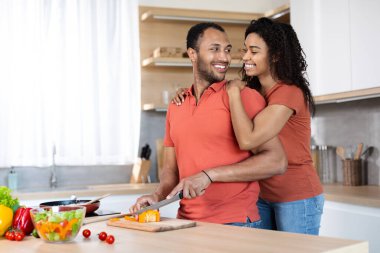 Happy young black couple in same t-shirts prepare homemade meal, wife hugs husband, man cuts vegetables in cozy kitchen interior. Love and relationships, healthy food at home and cooking together