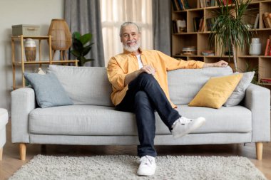 Relaxed grey-haired bearded senior man in stylish casual outfit sitting on grey couch at home, smiling at camera, cozy living room interior, copy space. Retirement, seniors lifestyle concept