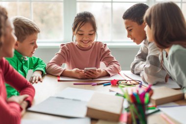 Diverse School Kids Using Cellphone During Break Having Fun Sitting At Desk In Classroom At School. Children And Gadgets. Educational Application On Smartphone. Selective Focus