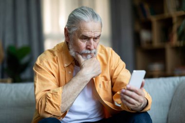Pensive elderly man sitting on couch at home, using smartphone, looking at gadget screen and touching his chin, having troubles with mobile application, copy space. Modern gadgets and senior people