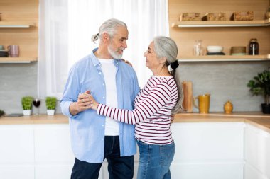 Portrait Of Romantic Senior Couple Dancing Together In Kitchen Interior, Happy Elderly Spouses Holding Hands And Looking At Each Other, Enjoying Spending Time With Each Other At Home, Copy Space
