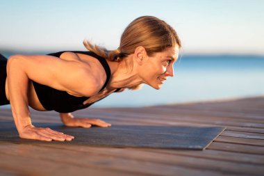 Closeup Of Athletic Middle Aged Woman Doing Plank Exercise Or Push Ups Outdoors, Sporty Female In Activewear Training On Wooden Pier Near River, Enjoying Outside Workouts, Side View With Free Space