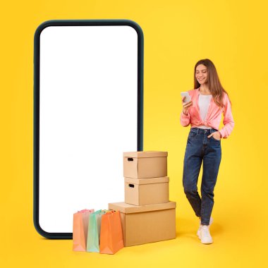 Delivery Application. Lady Standing Near Huge Cellphone, Shopping Bags And Cardboard Boxes Using Mobile Phone Buying Something Online Posing Over Yellow Background. Studio Shot, Square