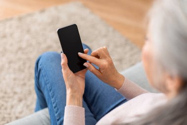 Caucasian mature gray-haired female typing on smartphone with empty screen in living room interior. Social media chat, new app, good website and technology for communication during covid-19 pandemic