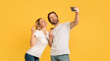 Cheerful excited young caucasian couple in white t-shirts have fun, make peace sign and selfie on phone, isolated on yellow background, panorama. Love, relationship photo for blog and social networks