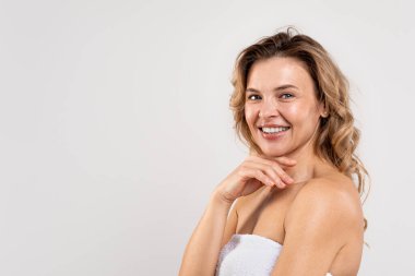 Mature Beauty Concept. Portrait Of Happy Middle Aged Woman With Beautiful Skin Standing Wrapped In Bath Towel Over Light Grey Background, Attractive Female Touching Chin And Smiling At Camera