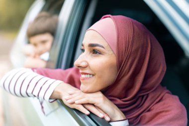 Smiling millennial arab female in hijab and boy look out the open window and enjoy journey in car and summer weekend, close up. Road to vacation with family, facial expression and life insurance