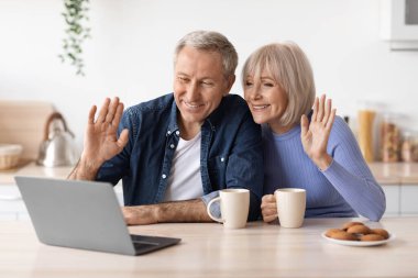 Cheerful elderly couple making video chat with friends while drinking coffee at kitchen, senior man and woman sitting at table, looking at computer screen, waving and smiling, embracing, copy space