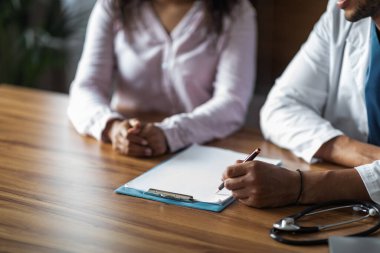 Cropped of black doctor and female patient sitting at table at clinic or hospital, having conversation, unrecognizable general practitioner taking notes at medical chart, copy space