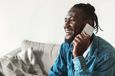 Cheerful African American Man Talking On Cellphone Having Pleasant Phone Conversation Sitting On Couch At Home, Side View. Mobile Communication, Technology And Gadgets Concept