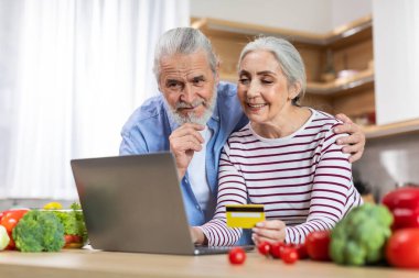 Happy Senior Couple Using Laptop And Credit Card In Kitchen Together, Smiling Elderly Spouses Ordering Groceries Or Food Delivery Online, Shopping In Internet Via Computer At Home, Closeup