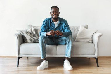 Successful Black Millennial Male Sitting On Couch Smiling To Camera In Modern Living Room Indoor. Man Posing At Home Enjoying Cozy Weekend. Front View Shot