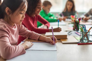 Korean Schoolgirl Writing Taking Notes Learning During Class Sitting With Multicultural Classmates In Classroom At School. Modern Education Concept. Selective Focus, Cropped