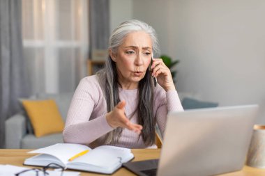 Sad serious caucasian elderly gray-haired woman speaks by phone and looks at laptop with client in living room interior. Problems at work at home, business with technology during covid-19 quarantine