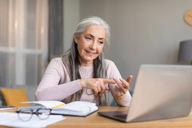 Glad friendly caucasian senior gray-haired female teacher speaks by laptop with student in living room interior. Video call, study, tutoring, work at home, business with tech due covid-19 quarantine
