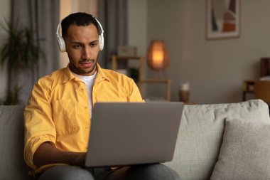Attractive young black man in headset using laptop computer, having online job, sitting on couch at home, blank space. Millennial African American male having video conference or business meeting