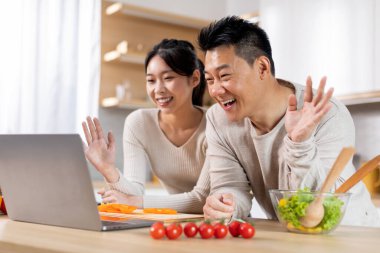 Happy asian couple having video chat with friends or family while cooking together at kitchen, standing next to table full of vegetables, waving and smiling at computer screen, copy space
