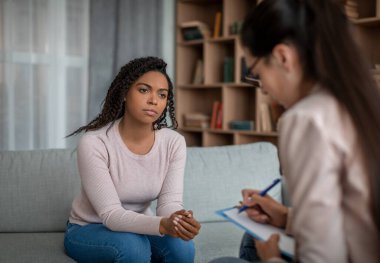 Personal mental consultation. Unhappy sad millennial black lady looking at european doctor woman in office clinic interior. Medical support, psychological therapy, session with professional and recipe