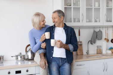 Cheerful elderly spouses enjoying time together at home, drinking coffee, happy senior lady sitting on kitchen table, hugging her handsome husband, couple having conversation, copy space