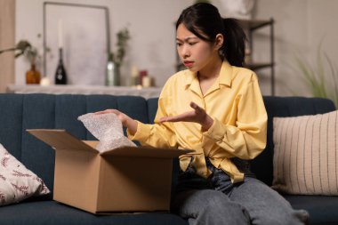 Discontented Asian Lady Buyer Unpacking Cardboard Box With Bad Product Sitting On Sofa At Home. Negative Feedback, Delivery Service And Shopping Problem Concept