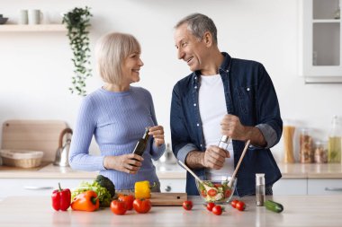 Beautiful senior spouses making healthy dinner together at home, happy elderly husband and wife cooking meal and having conversation, white cozy kitchen interior, copy space