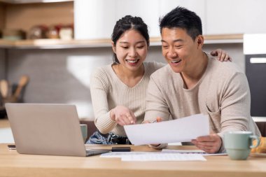 Cheerful asian spouses paying bills on Internet from home, sitting together at kitchen table, holding letters and papers, using computer and smiling, enjoying easy e-banking, copy space