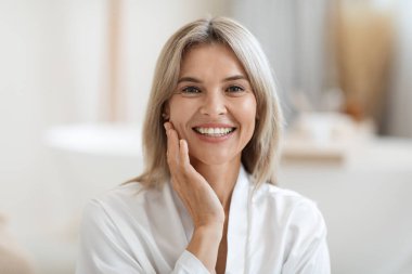 Closeup photo of attractive blonde middle aged woman in white silky bathrobe touching her pretty face and smiling, enjoying her morning face care routine, bathroom interior, copy space