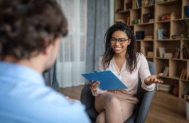Psychological therapy. Cheerful pretty young african american female psychologist in glasses talking with european man patient in office clinic interior. Personal consultation, depression treatment