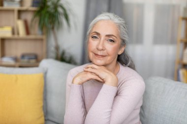 Smiling stylish confident attractive caucasian elderly gray-haired female looking at camera alone in living room interior, free space. Health care at home, mental balance, advertising and lifestyle