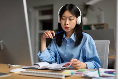 Korean Lady Designer Working With Color Samples Sitting Near Computer Wearing Headphones At Workplace In Modern Office. Young Graphic Designer Choosing Palette. Creative Career And Freelance