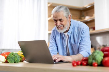 Portrait Of Smiling Senior Man Using Laptop In Kitchen, Happy Elderly Gentleman Typing On Computer, Searching Online Recipe Or Shopping In Internet While Cooking Lunch At Home, Free Space
