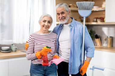 Domestic Chores Concept. Happy Senior Spouses Posing With Cleaning Supplies In Kitchen Interior, Beautiful Elderly Husband And Wife Smiling At Camera, Mature Couple Ready To Wash Home, Copy Space