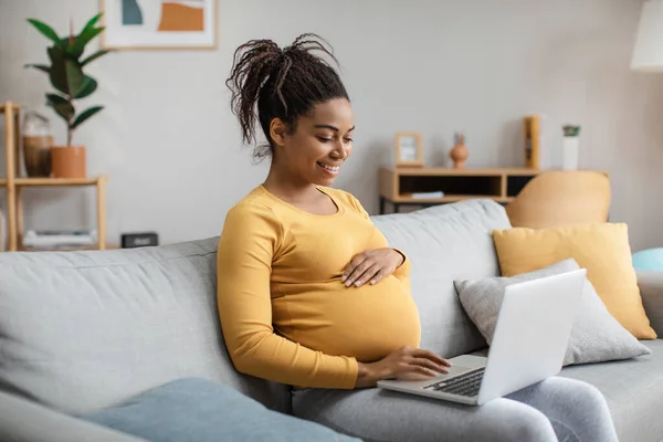 Smiling young pregnant african american woman with big belly working on laptop in living room interior. Motherhood, lady enjoy pregnancy at home, business and study in spare time, freelance and gadget