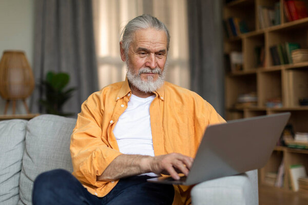 Handsome stylish grey-haired senior man websurfing while resting at home, sitting on couch and using modern computer, living room interior, looking at notebook screen and smiling, copy space