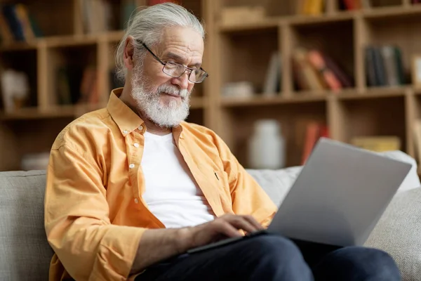 Old man in eyeglassses using modern laptop at home, handsome grandfather sitting on sofa with computer on his lap in living room, typing on notebook keyboard, sending emails, copy space